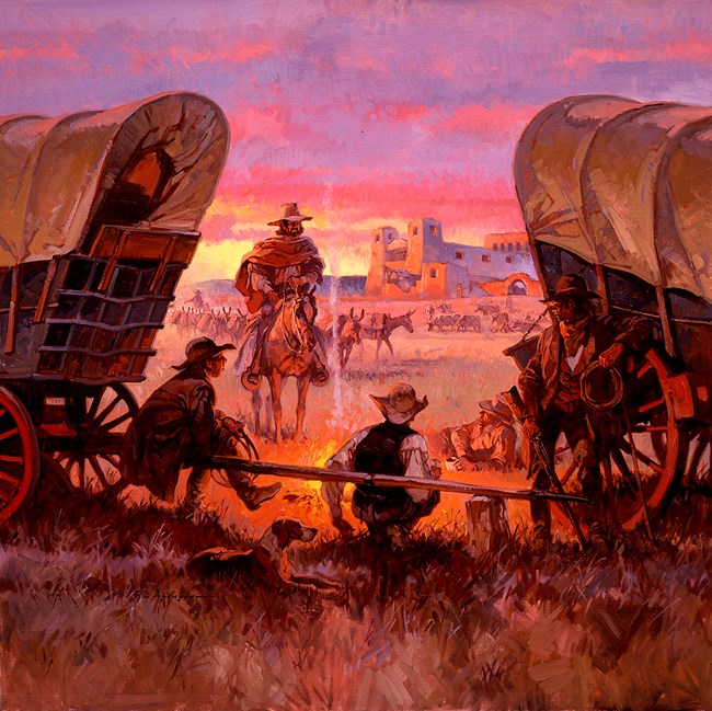 People gathered around a campfire with wagons on either side and a building in the background.