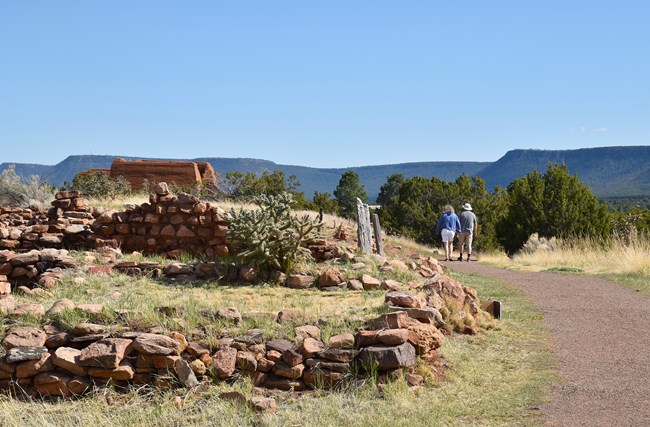 Two people walk near stone walls, flat green mesa top along the horizon.