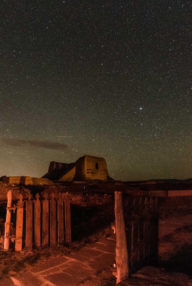 Gate in foreground, Pecos mission church with starry sky behind.