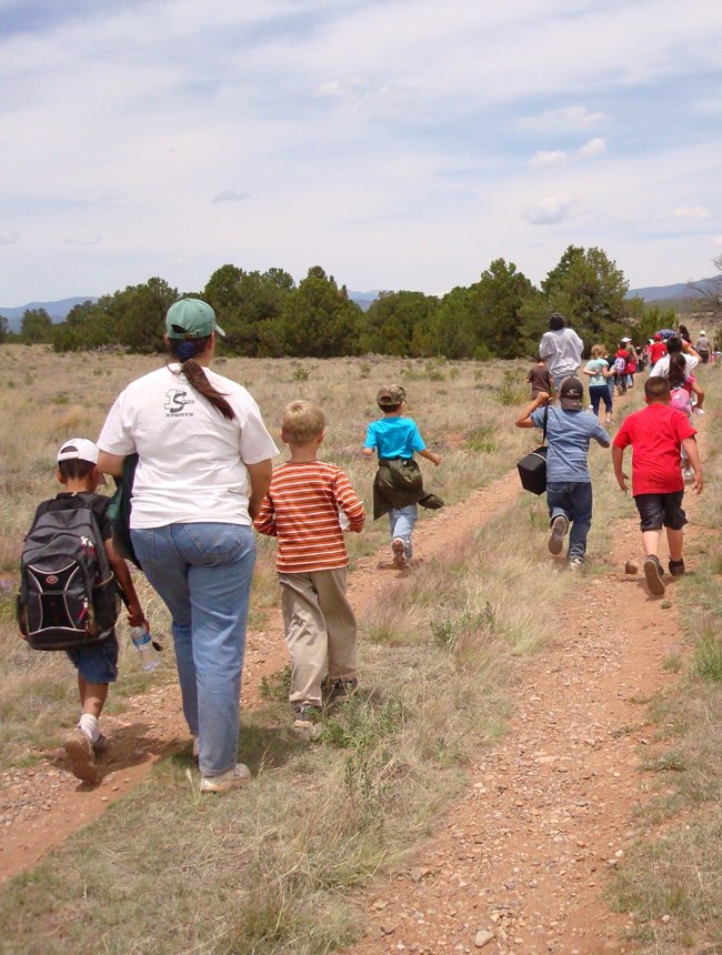 A large group of children run along dirt trail into the distance, with an adult chaperone walking at the end of the line.