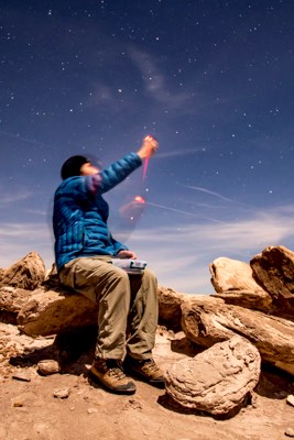 A man sitting on a boulder in the wilderness, holds a Unihedron sky quality meter up to the sky to determine darkness in the surrounding area.
