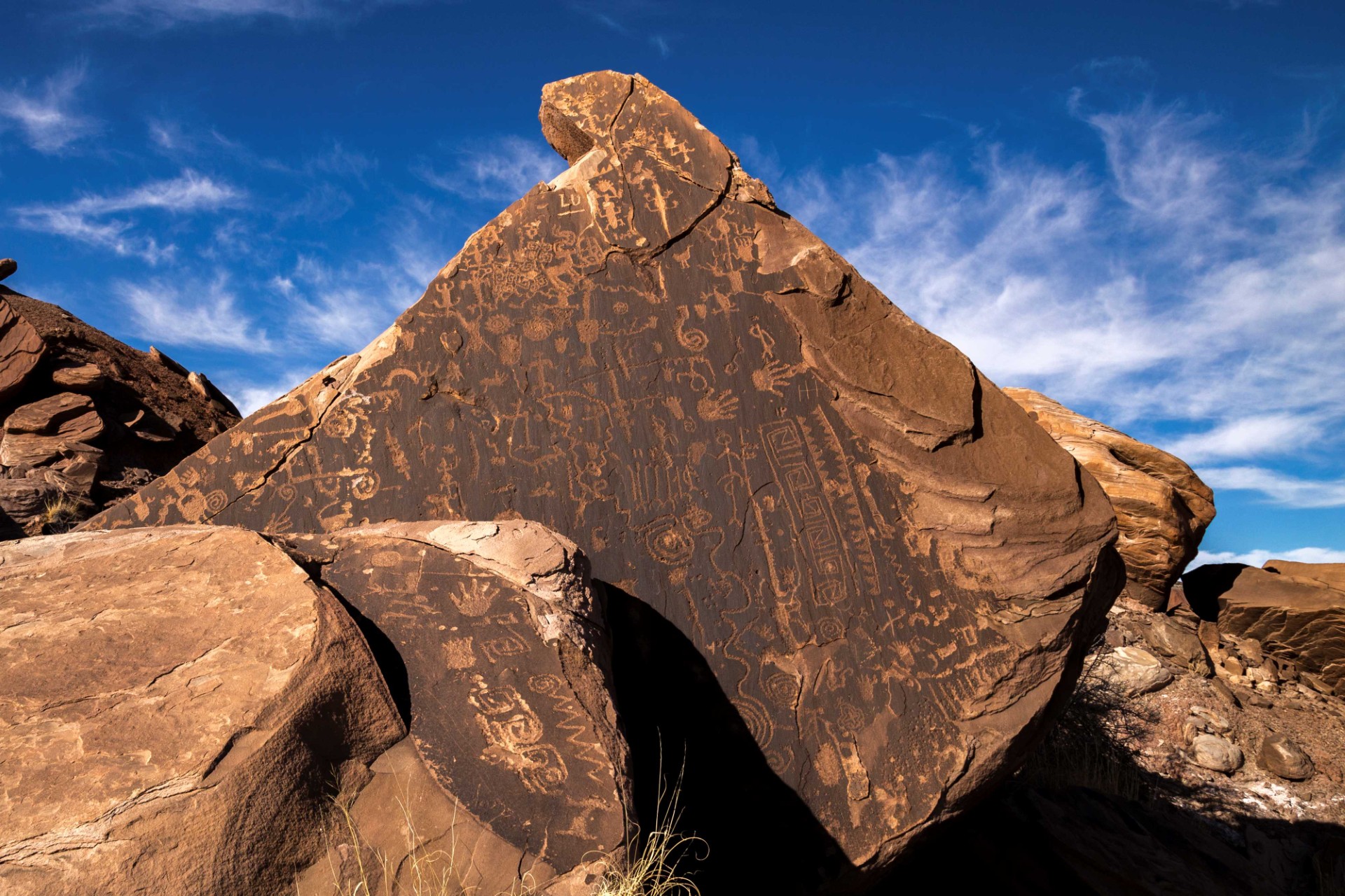 Flatiron Petroglyph Panel Pile of brown rocks with rock varnish, covered with many petroglyphs, under a blue sky with a few clouds.