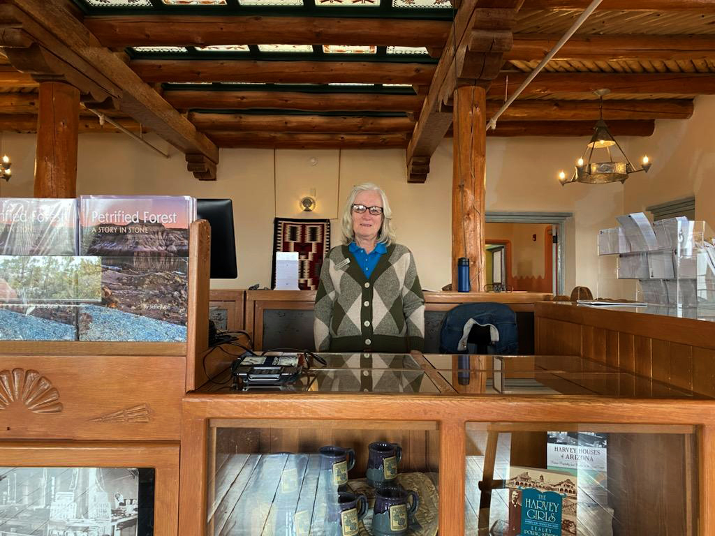 Museum Associate Debbie in the bullpen of the Painted Desert Inn National Historic Landmark: woman standing behind a counter in historic building with bookstore products.