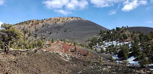 Volcanic cinder cone rises above cinder hills with trees and a blue sky above.