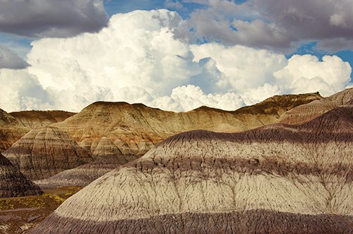 Puffy clouds over badlands with distinct banding in gray, purple, and blue