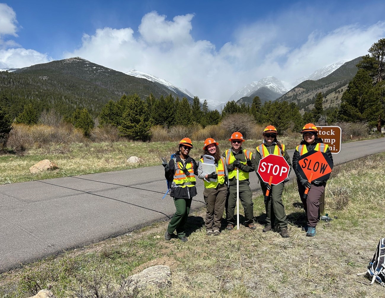 Trail Monitoring Team A group of five individuals wearing hard hats, and orange vests stand in a grassy field with a background of mountains. They are holding stop signs.