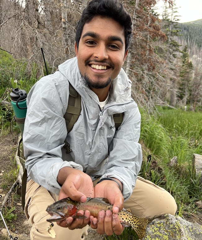 Prineet holding a fish A young man kneals down on the ground smiling holding a fish in both hands