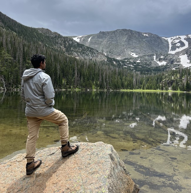 Prineet hiking A young man stands on a large rock facing a body of water and mountains