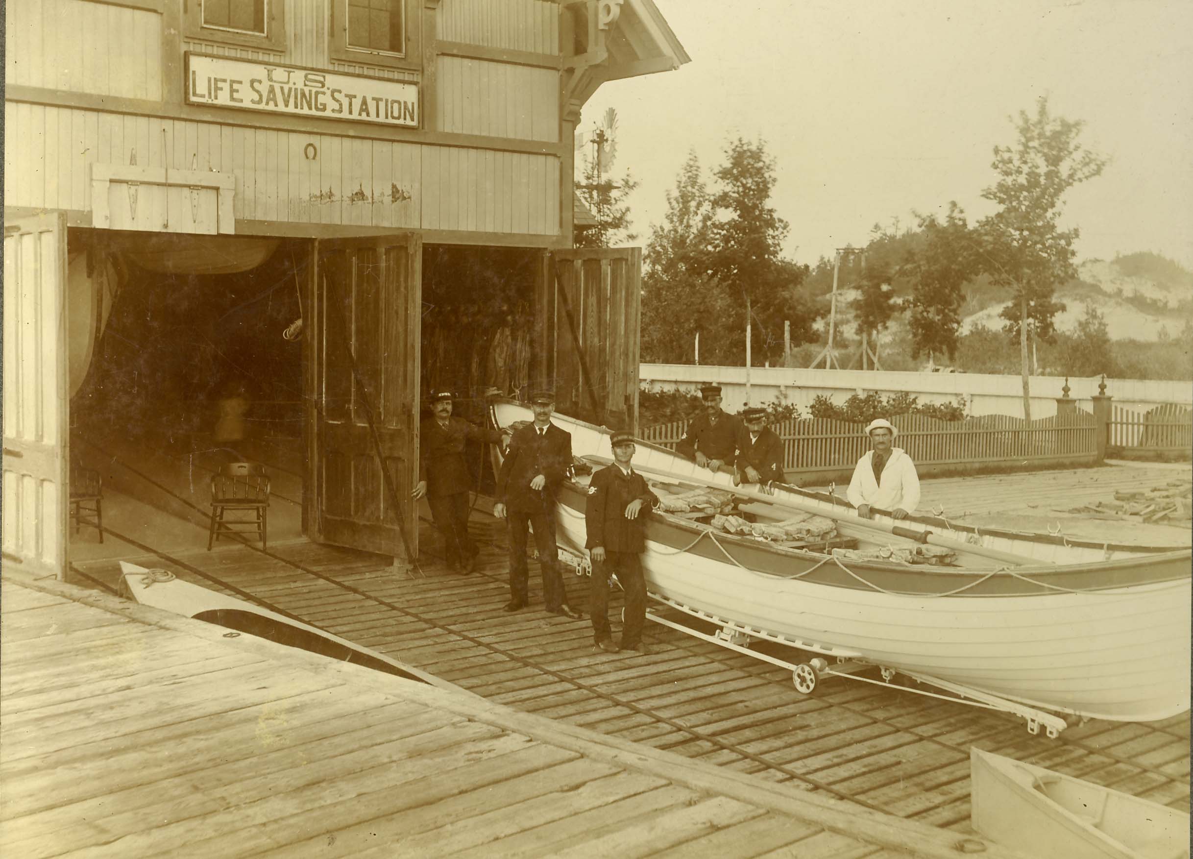 historic image of men in uniform standing next to a surfboat in front of a building with two large doors. Sign above doors reads US Life Saving Station.
