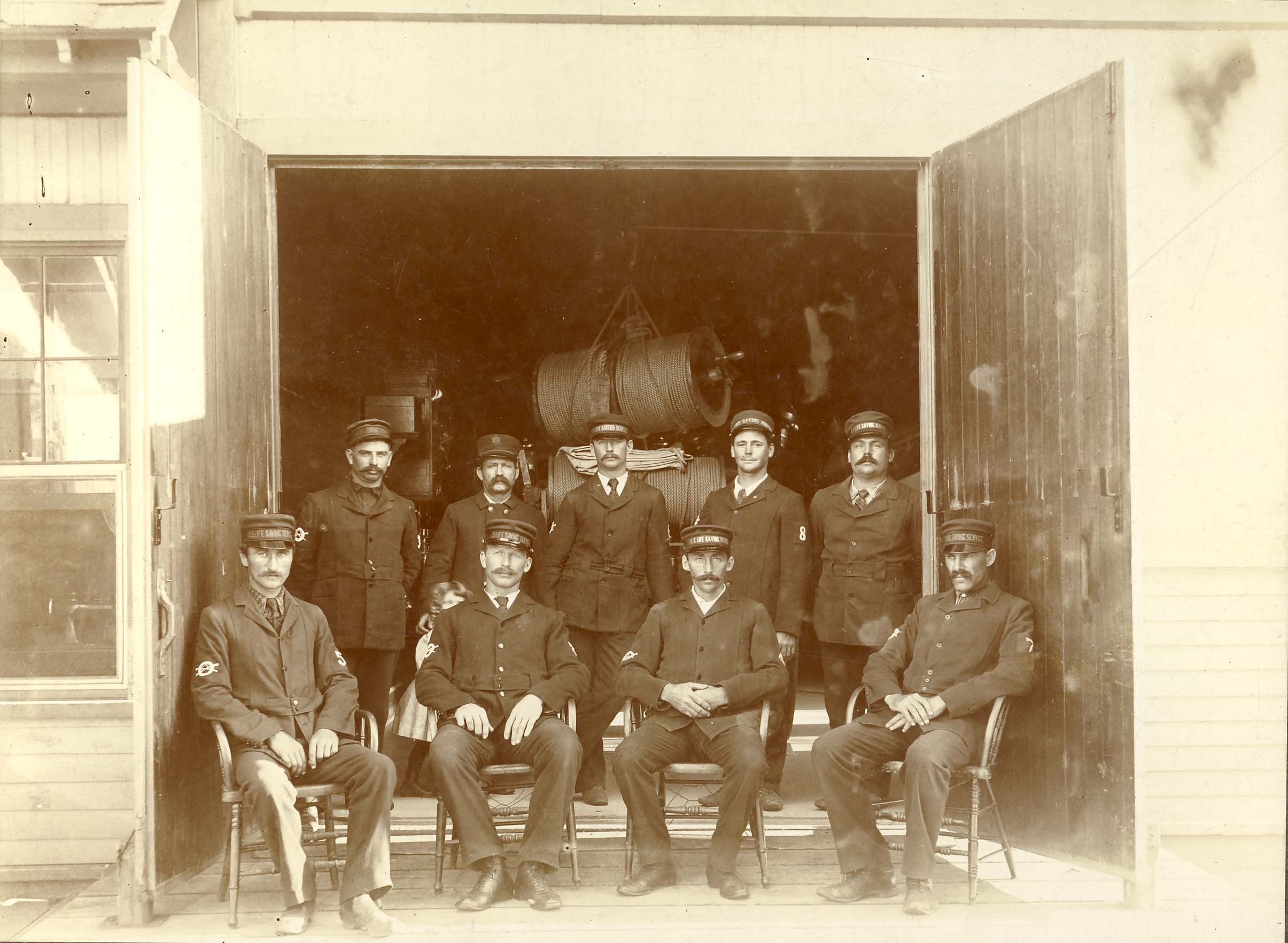 9 men in US Life Saving Service uniforms pose for a group photo inf front of a building with large open door.