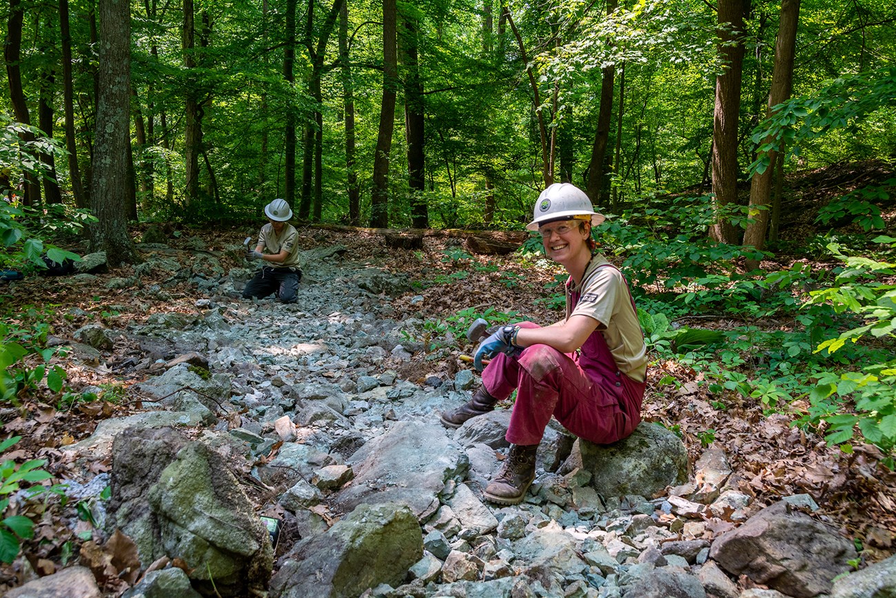 Trail crew on rocks A young woman wearing brown pants and red pants sits on a rock among a field of rocks. Another individual sits in the back sitting on rocks