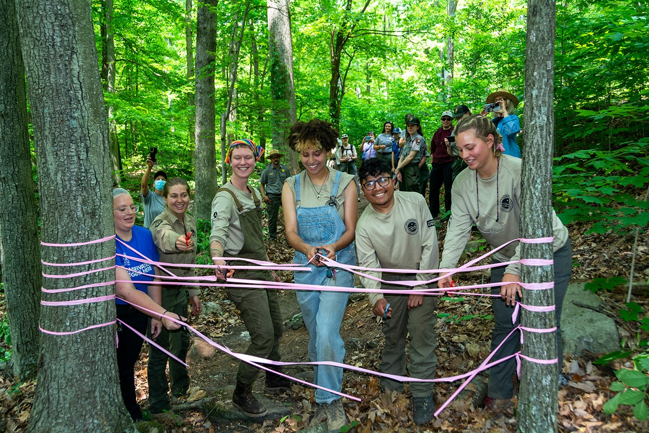 Trail Crew Ribbon cutting A group of six youth wearing shirts that say Conservation Legacy and SCA stand in the woods surrounded by other individuals. They are cutting a pink ribbon connected between two trees with scissors.