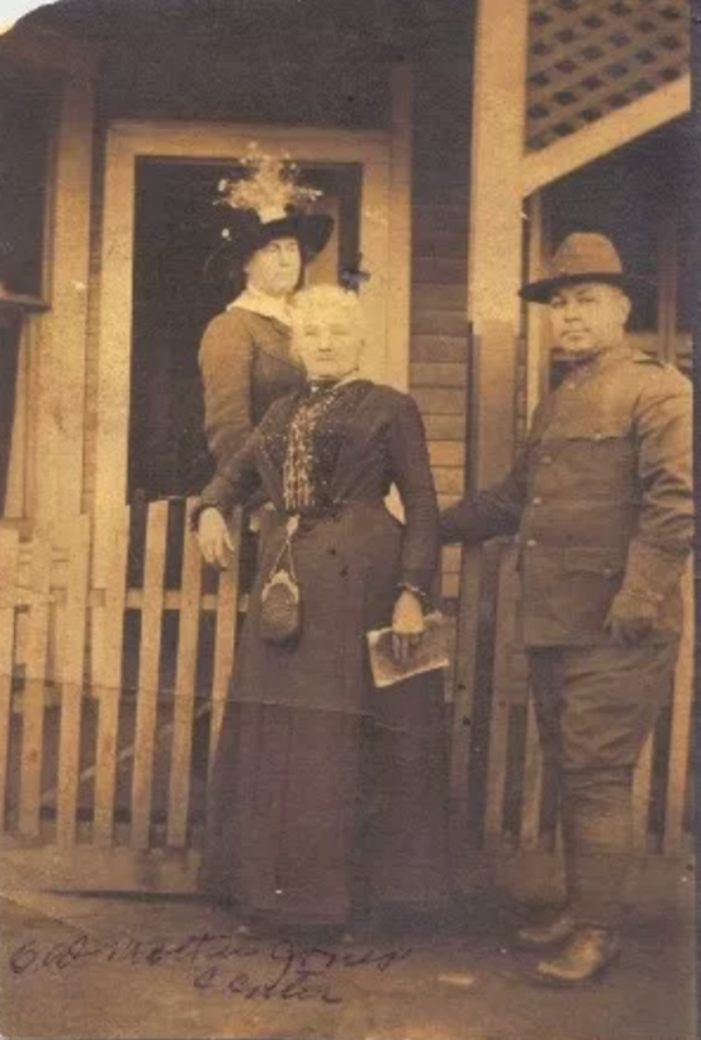 Three people are posed in front of a small house, one woman behind a fence, with another woman and a man in front of the fence.
