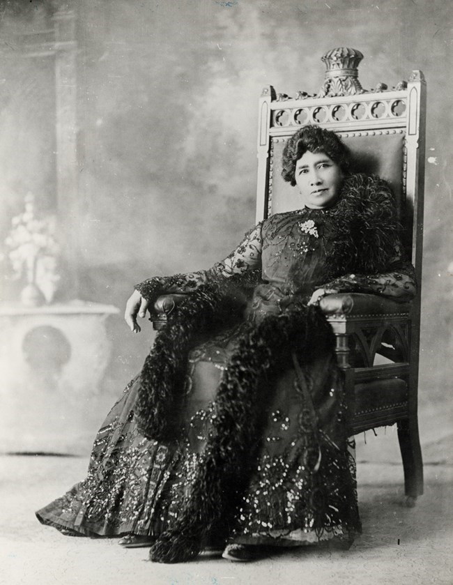 A black and white image of Queen Lili‘uokalani in a dark dress, sitting in a chair and staring at the camera.