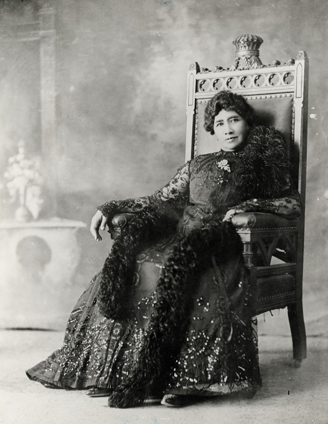 ALKA_Lili‘uokalani A black and white image of Queen Lili‘uokalani in a dark dress, sitting in a chair and staring at the camera.