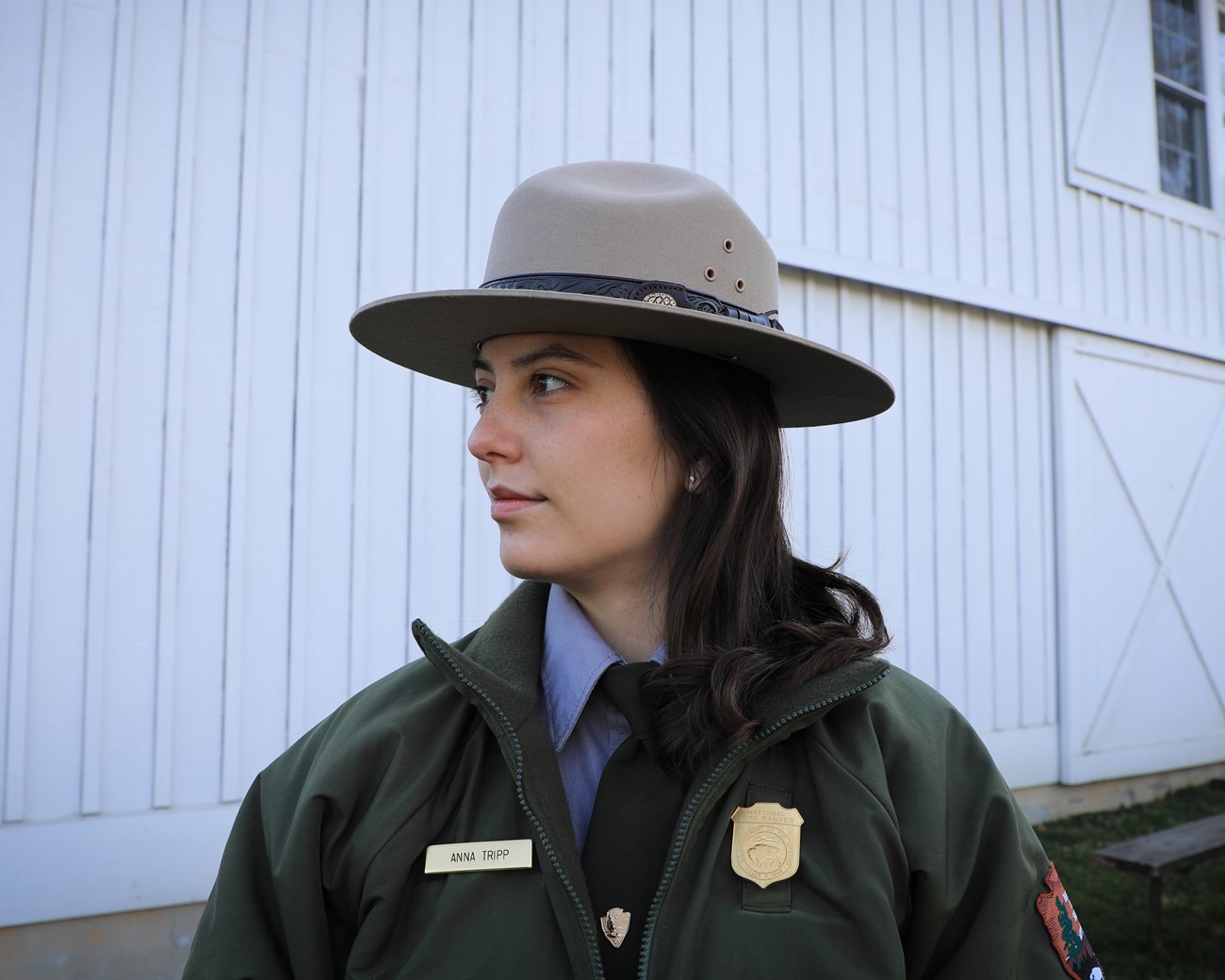 AnnaTripp_WinterHeadShot_NPS A white female National Park Service ranger poses looking off to the left in front of a white barn, part of a picnic table can be seen to her right.
