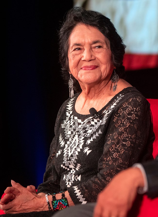 A colored photo of Dolores Huerta sitting down with her hands together. She wears a black and white styled shirt, and smiles at the person sitting next to her.