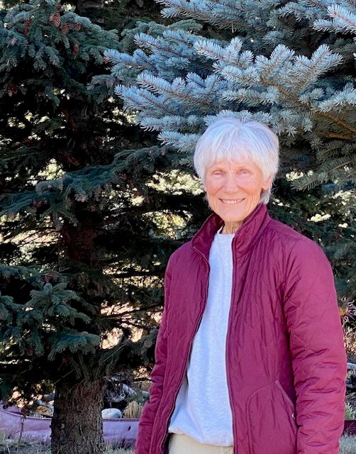 A photo of Elizabeth Horn wearing a white shirt and pink jacket, smiling for the camera in front of green trees.