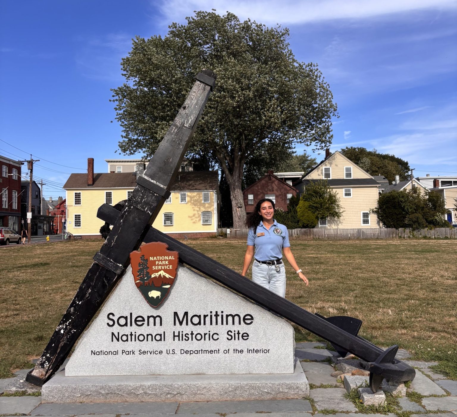 Young woman wearing a light blue shirt stands in front of a cement sign that reads “Salem Maritime National Historic Site; National Park Service U.S. Department of the Interior” in black lettering. There is a large black anchor resting diagonally on the