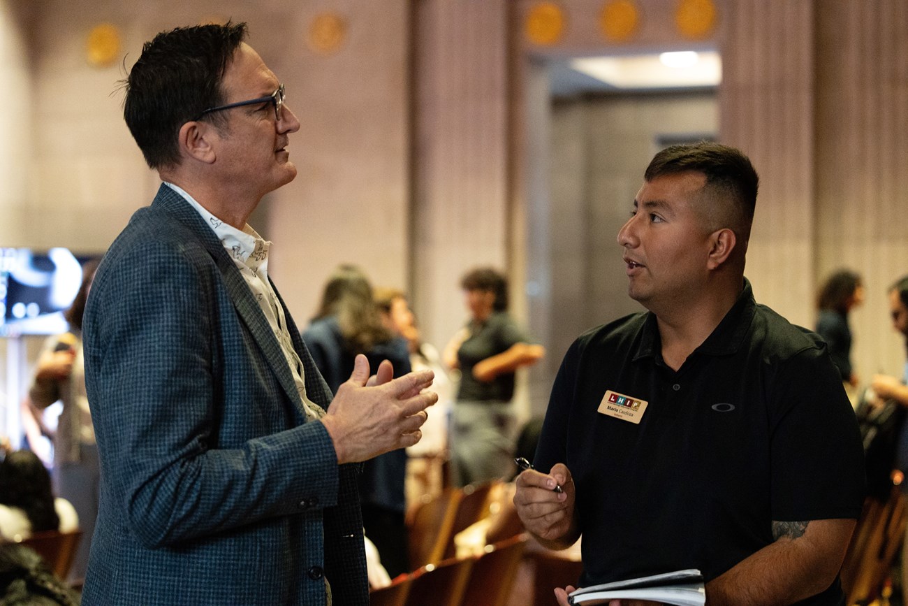 MarioTalking Two men, one wearing a suit, the other wearing a polo, stand facing each other in conversation