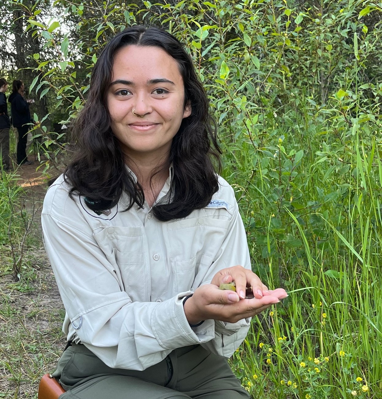 A young woman kneels in the grass holding an insect in her hands