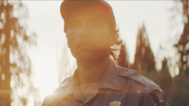 A close-up of a man, Garrett Dickman, in a National Park Service uniform and a baseball cap. There is an orange glow to the photo, indicating a sunset behind him.
