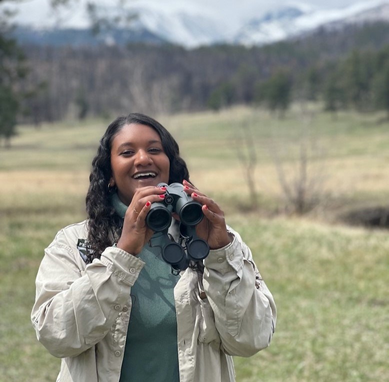 A young woman wearing a beige jacket outside while holding binoculars