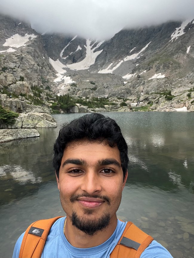 Prineet by a Mountain A close up of a young man standing outside in front of a vast landscape with mountains, clouds, and a lake