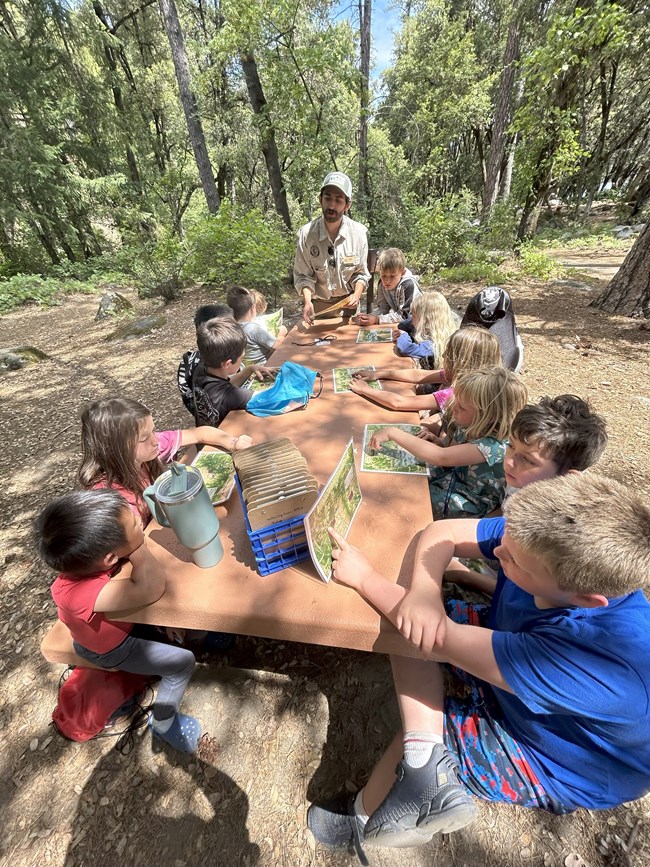 A young man stands at the head of a picnic table surrounded by children. They all have activity sheets/maps in front of them