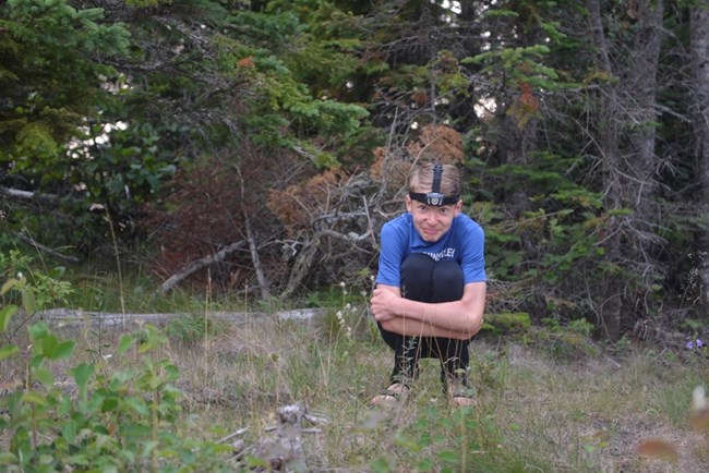 sandaled child in a squatting position in the woods with headlight on head and at the ready even though the daylight is abundant