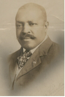 Portrait of bald black man with dark mustache and wearing a suit jacket with vest and a tie.
