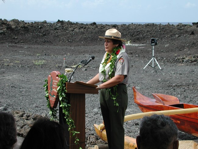 A photo of Geri Bell at the Kaloko-Honokōhau National Historic Park visitor center, speaking at a dedication ceremony outside and in front of a crowd.