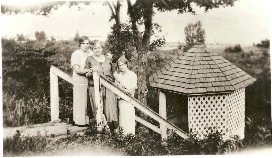 An adult and two children stand outside a grassy area. The wooden steps they are on lead down to a wood latticed well-gazebo.