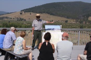 Male in ranger uniform wearing dark glasses gives outdoor presentation to visitors facing him.