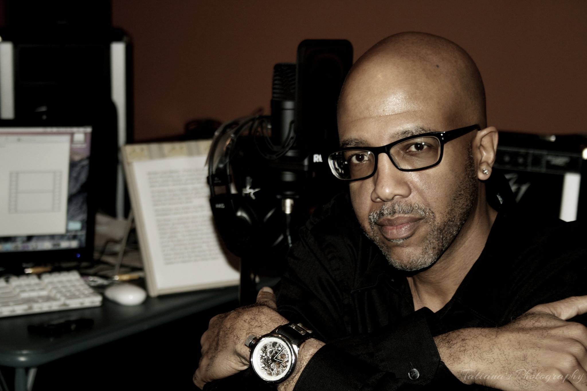 A man sits in a black shirt and with crossed arms at his computer and recording gear on the desk behind him.  He wears glasses and looks at the camera.
