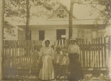 Black and white image of an African American woman standing in front of a house with three children.