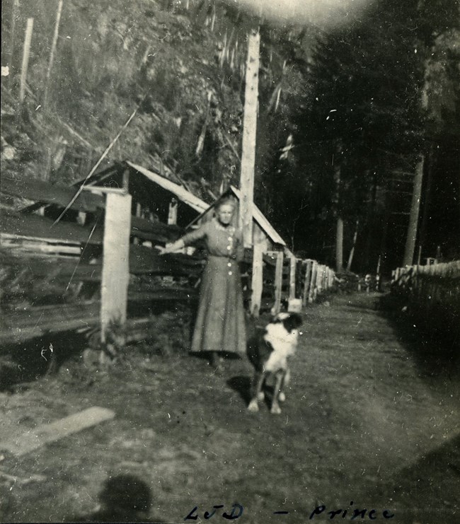 An old photo of Lucinda Davis leaning against a fence at the family homestead with her dog to the left of her.