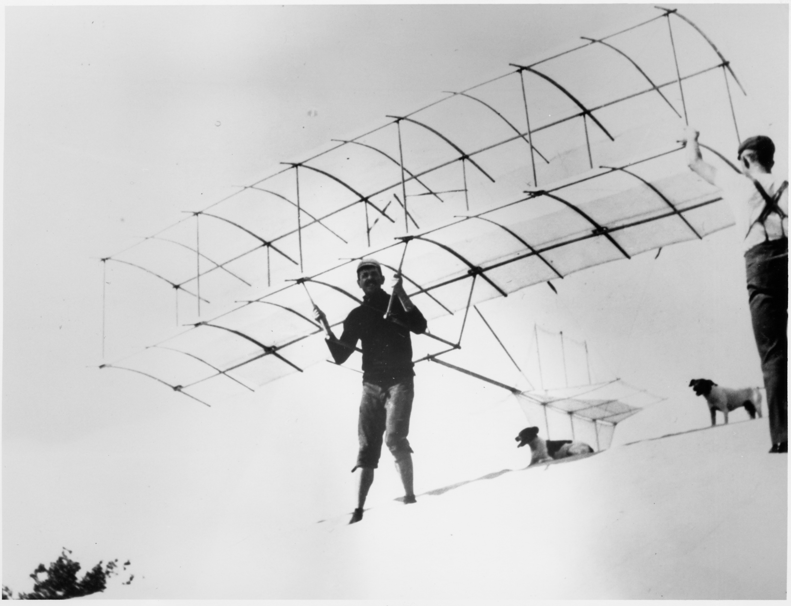 historic image of a man about standing on a hill about to take off in a glider from the standing position. A man and two dogs are also in the photo. 
