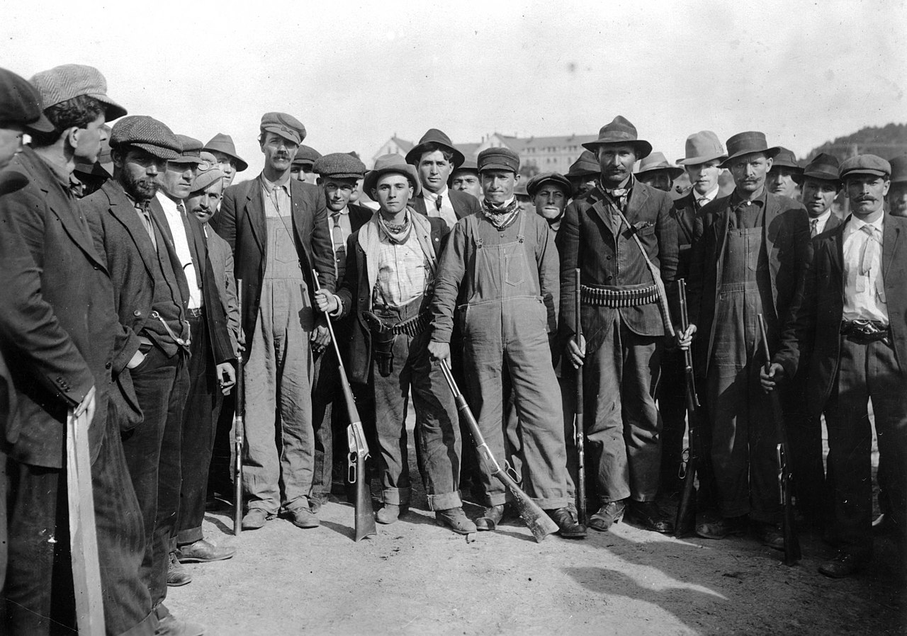United Mine Workers 25 United Mine Workers pose for a picture outside. They are dressed in civilian clothes, like overalls, and are shown holding rifles and wearing ammunition belts.