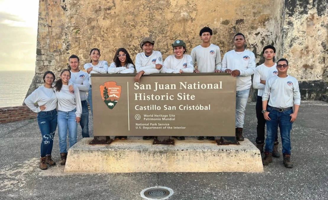 San Juan A group of nine individuals standing outside in front of a sign that reads "San Juan National Historic Site: Castillo San Cristobal"
