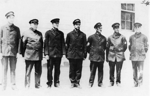 7 men in US Life Saving Service uniforms pose in front of a white building.
