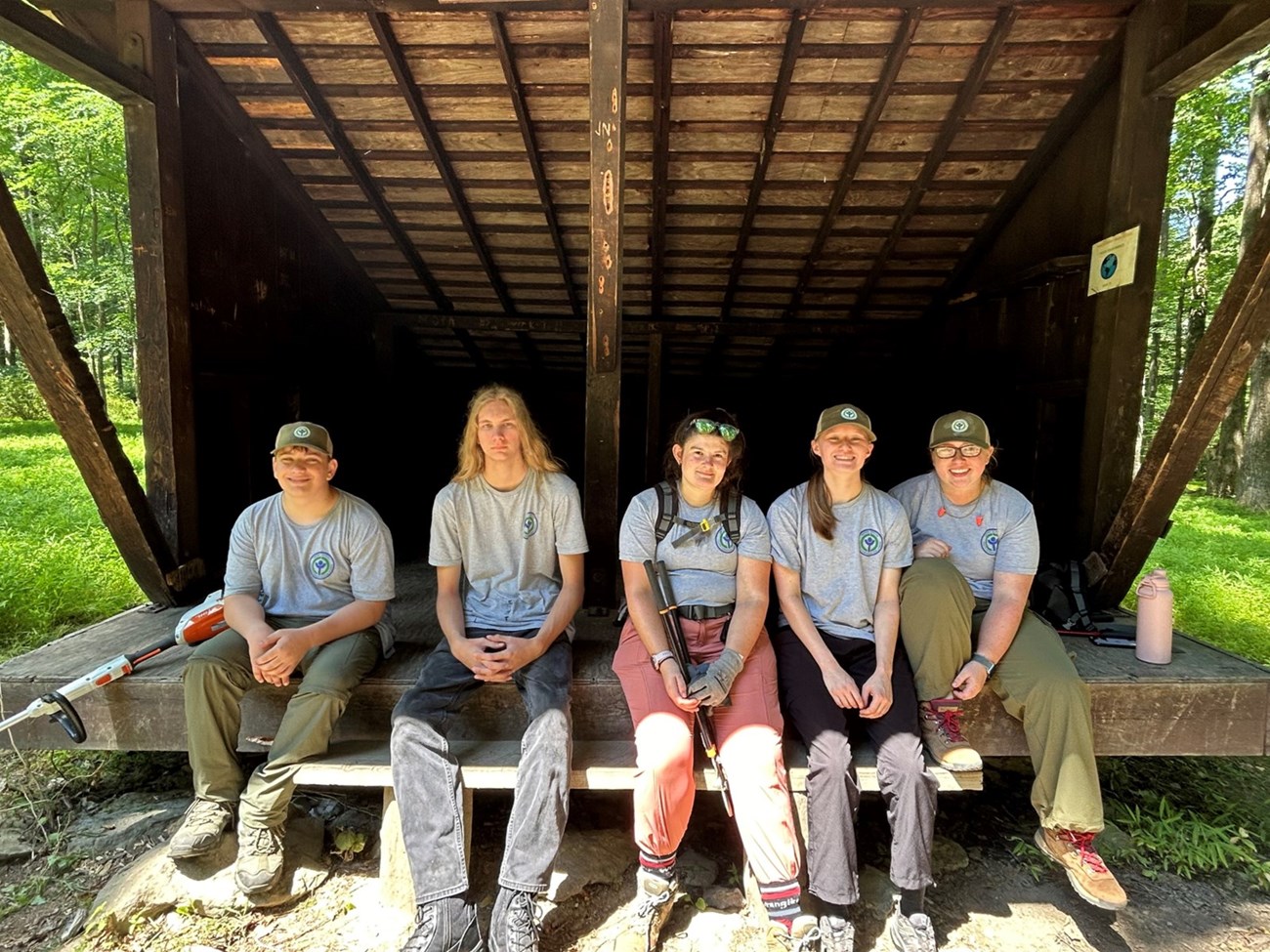 YCC crew Five youth wearing hats sit outside under a shed structure. There are two mowing devices next to them.