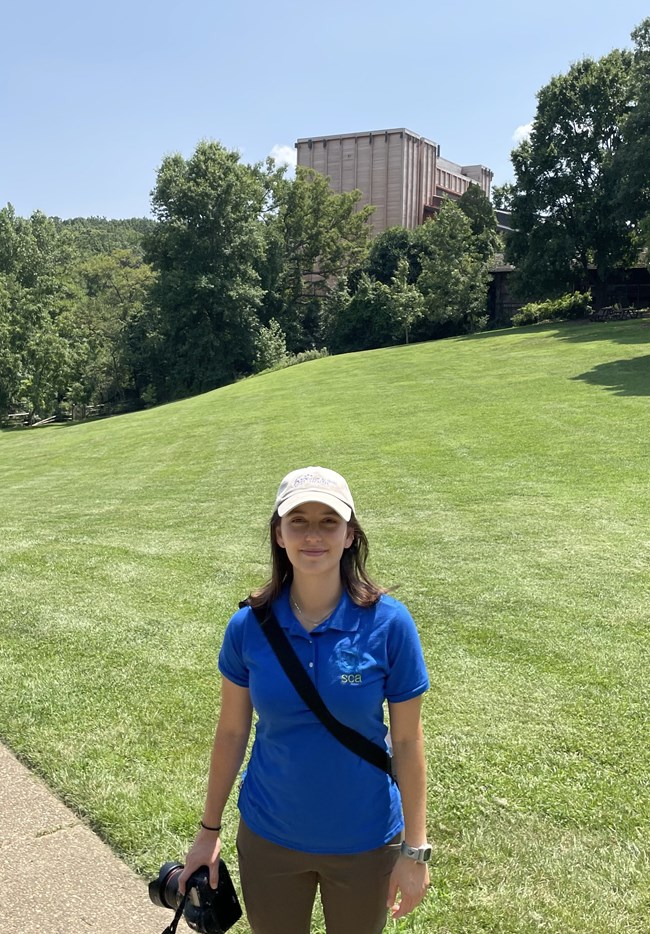 SCA intern A white female stands in front of an open grassy area, with a wooden amphitheater behind trees and a camera in her hand. She’s wearing a blue polo with a logo that reads “SCA”.