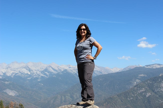 Caro Luévanos-Garcia smiling for the camera as she stands on a Moro Rock in front of a blue sky and mountain range.