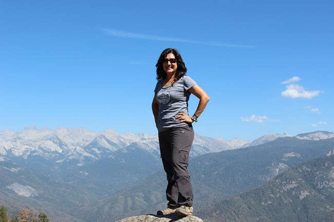 SEKI_Caro Luévanos-Garcia Caro Luévanos-Garcia smiling for the camera as she stands on a Moro Rock in front of a blue sky and mountain range.