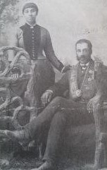 Sarah in a beautiful dress stands next to William who wears and ornate suit and sits in a chair.