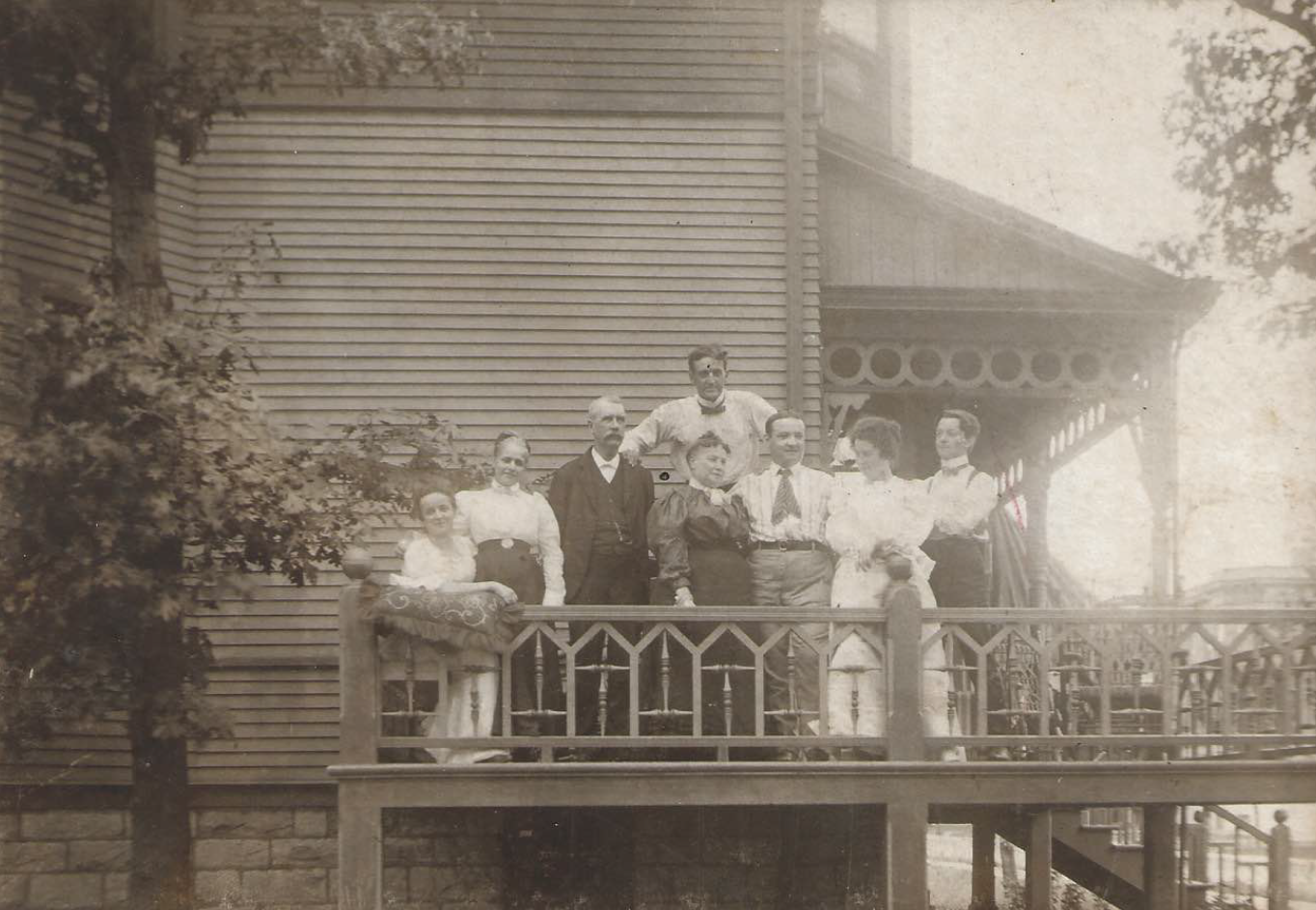 A family is gathered on the balcony of a large cottage with smiles on their faces. One family member is tall above the everyone else, one sits, and the rest stand on the balcony floor.