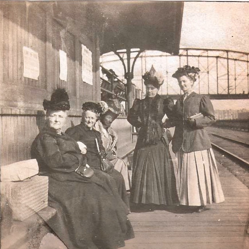Five people wait at a train station, all well dressed and wearing elaborate hats. Three of them sit while two stand.