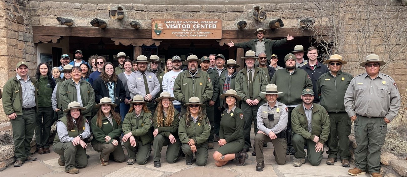 A large group of rangers in green and grey stand in front of the Bandelier Visitor Center pueblo revival style building.
