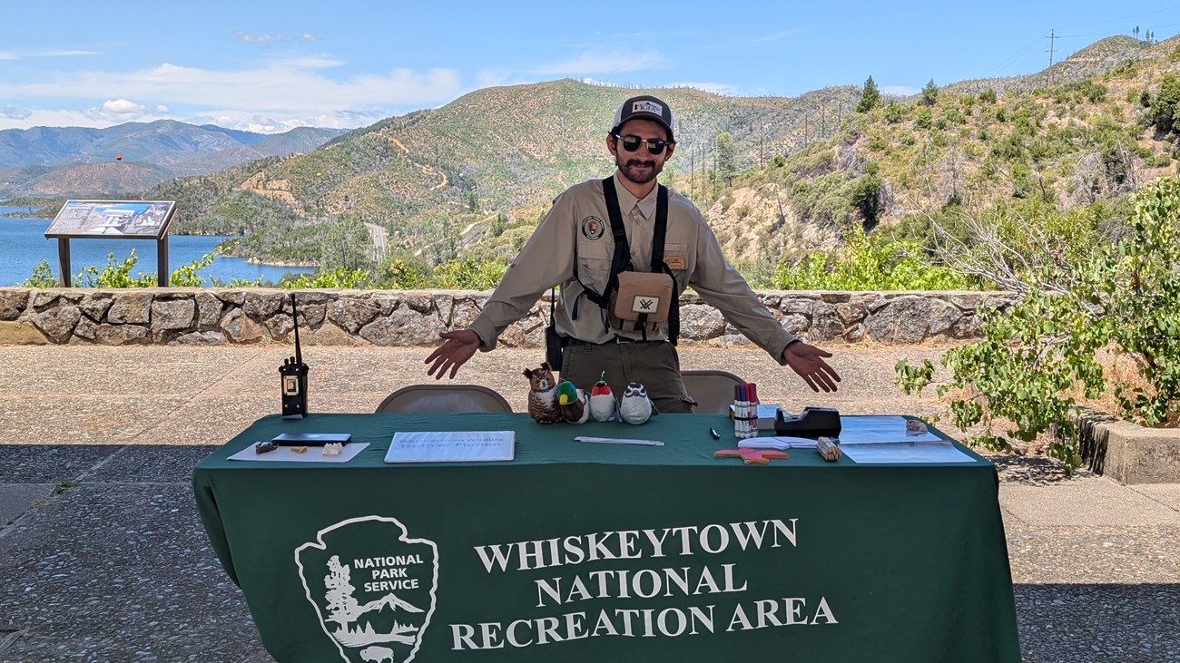 A young man stands behind a green table with supplies on it. In the background there is a mountain landscape.
